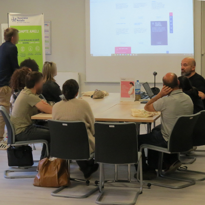 Futurs parents assis autour d’une table regardant un diaporama projeté et expliqué par un collaborateur de la CPAM de Charente-Maritime, dans une salle de réunion.