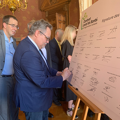 Laurent FLAMENT, directeur de la délégation département de l’ARS Nouvelle-Aquitaine et David XARDEL, directeur de la caisse d’assurance maladie de la Charente-Maritime entrain de signer le contrat local de santé en grand format disposé sur un chevalet, dans une salle de la Mairie de La Rochelle.