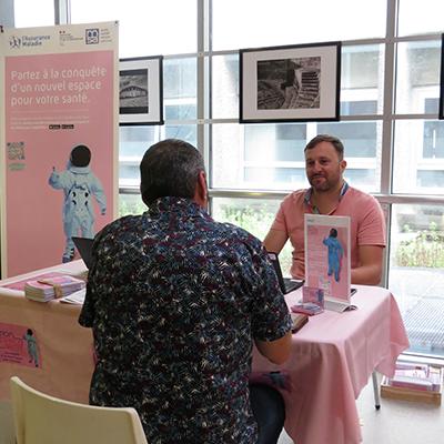 Stand d’information sur le dispositif MES avec un collaborateur de la CPAM recevant un assuré, de dos, assis autour d’une table, dans le hall de l’hôpital de La Rochelle.