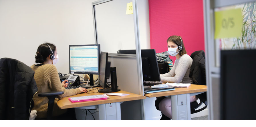 photo de 2 agents de la CPAM à leur bureau, le casque téléphonique sur la tête, face à leurs ordinateurs.