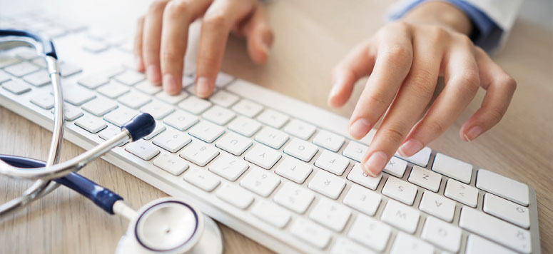 photo des mains d'un médecin sur son clavier, avec un stéthoscope posé sur son bureau.