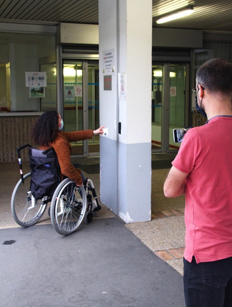 Photo du tournage de la vidéo des élèves du Lycée Joseph Roussel dans les locaux de la CPAM de la Sarthe : une jeune femme en fauteuil roulant passe son badge pour entrer dans le bâtiment.